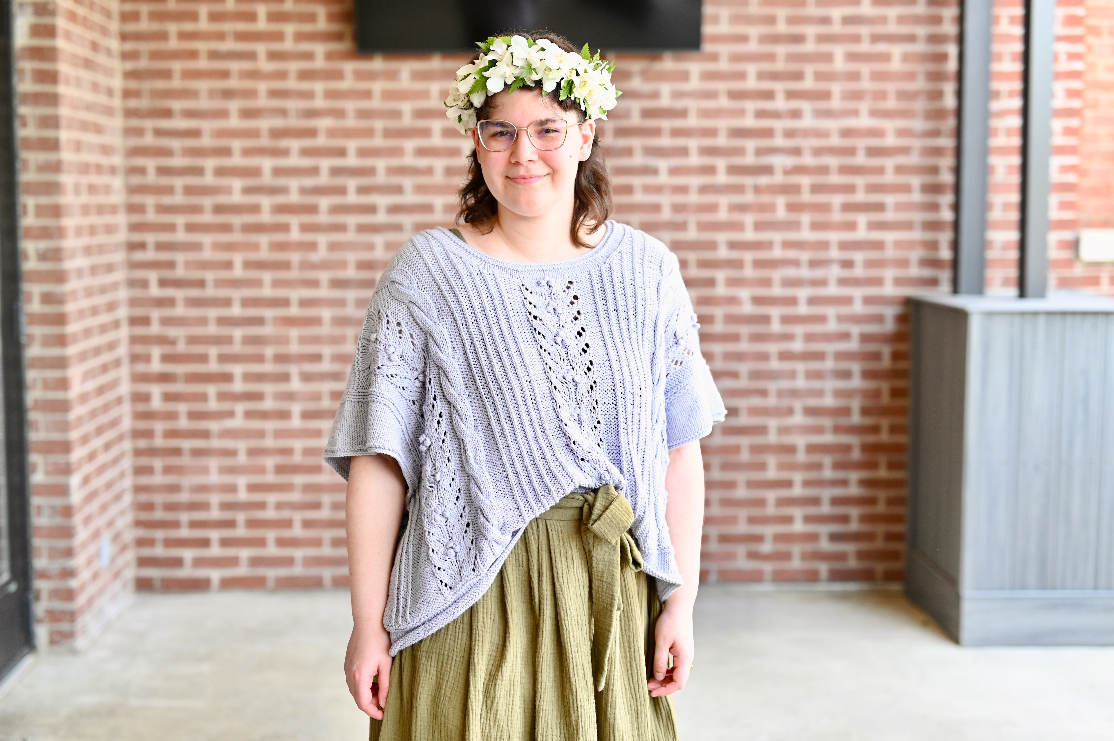 Frankie Knuckles, a white non-binary person with dark hair, wearing glasses, a Lei Po'o, a light purple handknit shirt, and a dark green skirt.